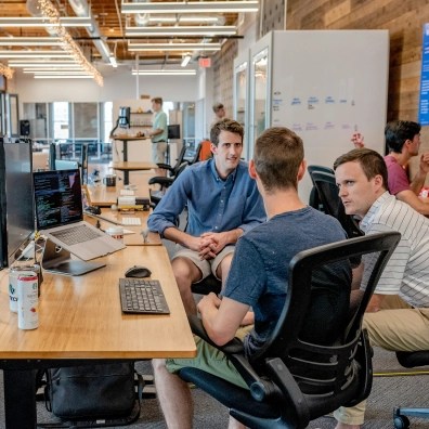 A group of three males sitting around a desk, talking to each other.