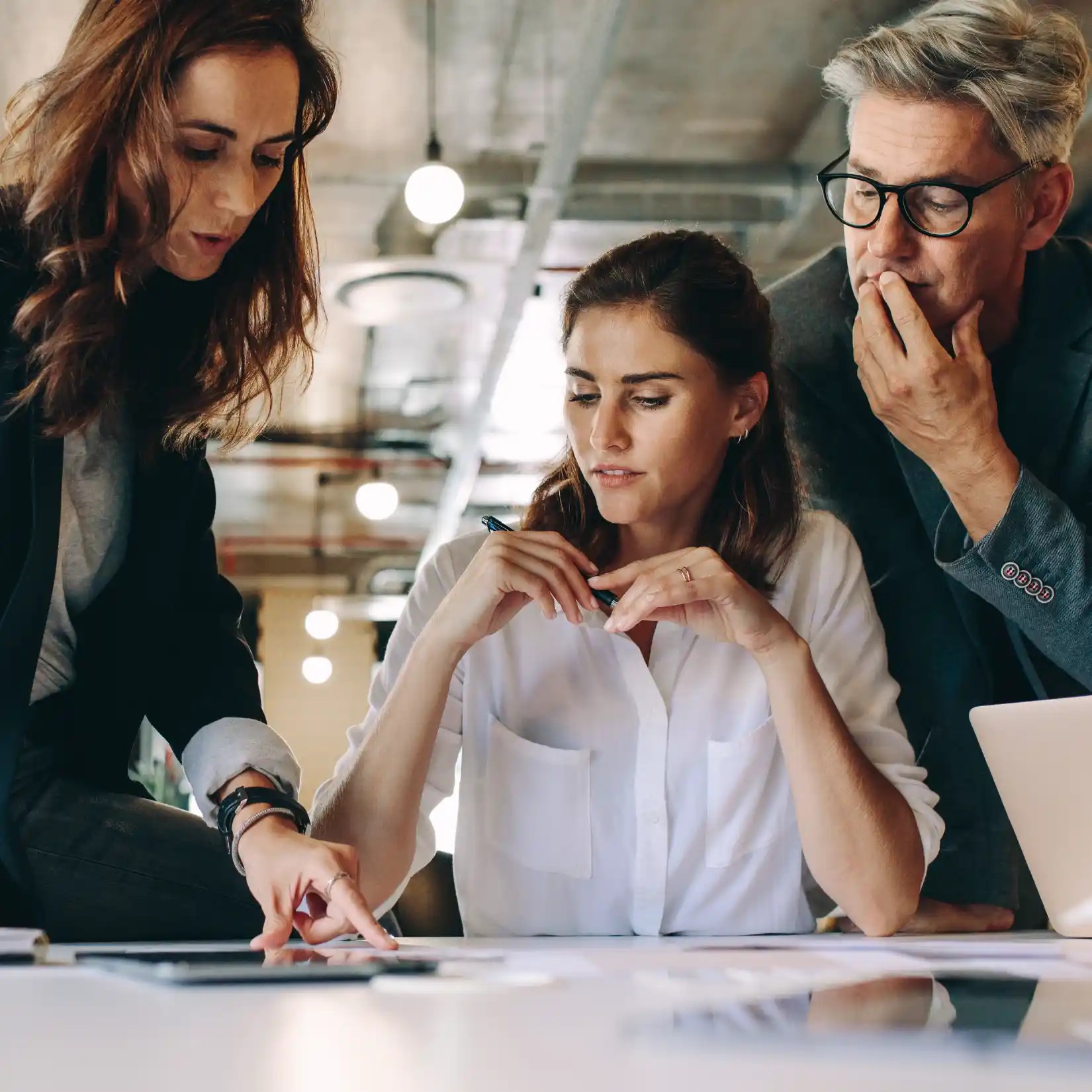 Three colleagues in a modern office closely reviewing documents and a tablet during a team discussion