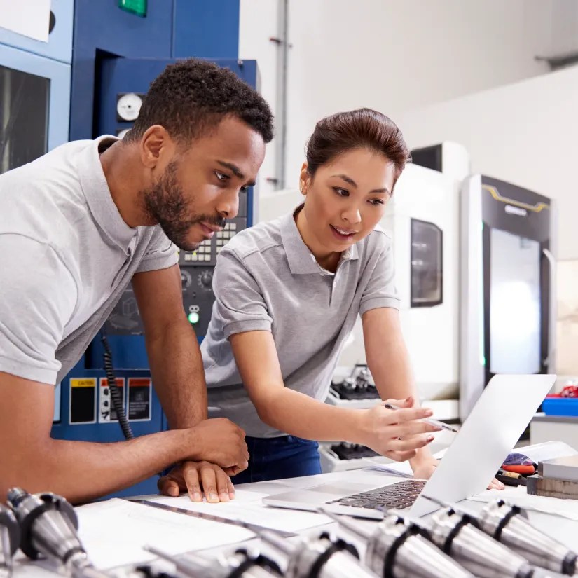 Two engineers reviewing plans on a laptop inside a manufacturing facility."