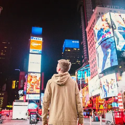 Person in a hoodie staring up at digital billboards in Times Square at night